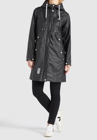 Černý voděodolný plášť do deště s kapucí, předním zipem a patentkami; obsahuje bílé šňůrky a boční kapsy. Nosený přes černé legíny a tenisky.