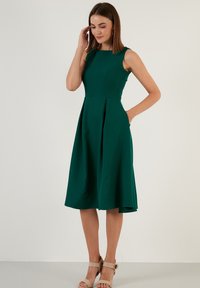 Rochie verde, fără mâneci, cu un corset ajustat și fustă evazată; are buzunare laterale și un tiv până la genunchi. Material neted cu o textură moale.