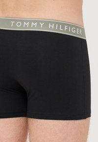 Close-up de uma pessoa a usar roupa interior boxer preta com uma cintura cinzenta que apresenta a marca "TOMMY HILFIGER".