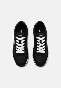 Baskets de sport noires avec des lacets et un logo blancs, présentant une texture lisse et une semelle blanche contrastante. Design à bout rond, semelle intérieure rembourrée.