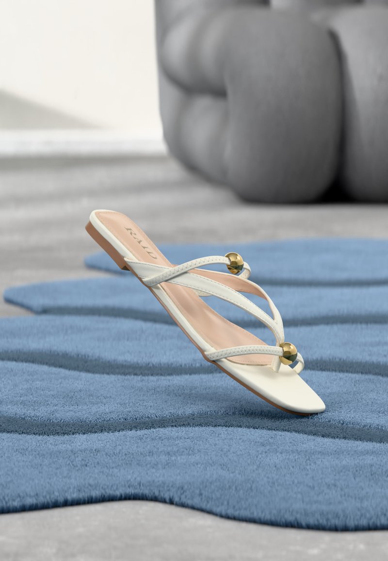 Sandale blanche à bout ouvert avec fines brides croisées et accents de boules dorées, présentée sur une surface texturée bleue.