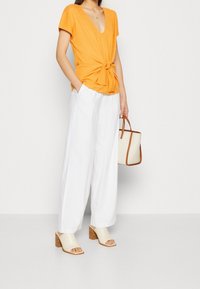 Top naranja con escote en V y detalle anudado, pantalones anchos blancos y sandalias de tacón beige; llevando un pequeño bolso crema con detalles en marrón.