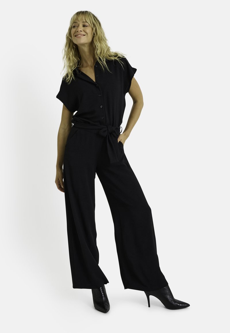 Mono negro con mangas cortas, cierre de botones y cintura con lazo. Los pantalones son de pierna ancha y se combinan con tacones negros de punta.