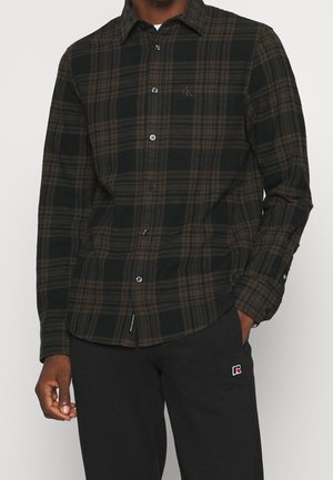 Homme portant une chemise à carreaux noire et brune avec poche poitrine et un pantalon noir avec un petit logo sur la cuisse gauche.