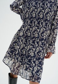 Robe bleu marine avec un imprimé floral, fabriquée en tissu léger et transparent. Présente des manches longues, une texture plissée et un ourlet à la cheville.