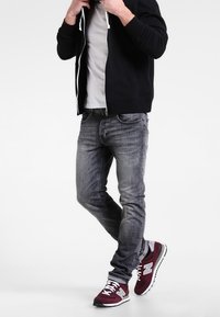 Persona vistiendo una sudadera con cremallera negra, una camiseta gris claro, jeans gris desgastados y zapatillas maroon de New Balance, de pie con una pierna cruzada.