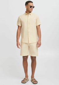 Chemise beige clair à manches courtes et shorts assortis, en tissu texturé, associés à des sandales brunes et des lunettes de soleil noires.