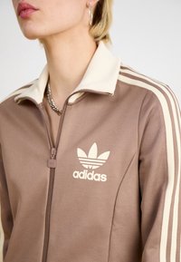 Brun zip-up träningsoverall med krämfärgade detaljer, med Adidas-logotyp och tre vita ränder längs ärmarna, tillverkad av ett slätt tyg.