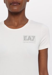 Balts pieguļošs T-krekls, izgatavots no mīksta kokvilnas, ar dekoratīvu sudraba rhinestone logo uzlīmi krūšu daļā ar tekstu "EA7" un "Emporio Armani".