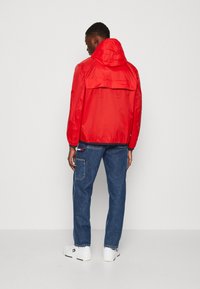 Rød vindjakke med hætte, elastiske manchetter og rygventilation, parret med blå denimjeans og hvide sneakers. Glat, blank tekstur.