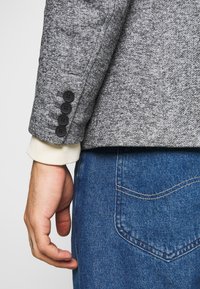 Grau strukturierter Blazer mit vier schwarzen Knöpfen am Ärmelaufschlag, kombiniert mit einem cremefarbenen Innenfutter und blauen Jeans.