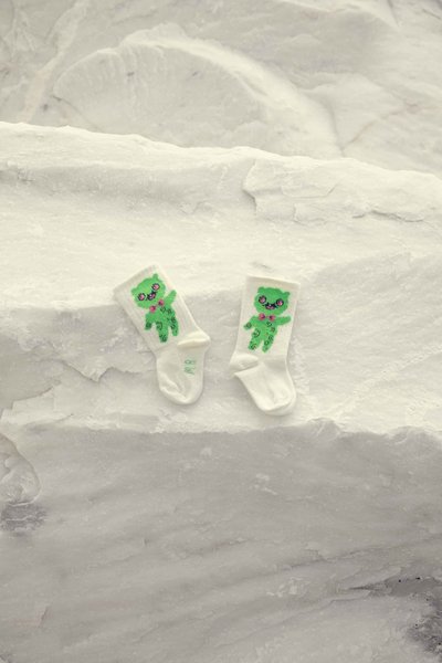Ζευγάρι λευκών παιδικών καλτσών με ένα χαμογελαστό πράσινο αρκουδάκι που φορά ροζ παπιγιόν, τοποθετημένα σε υφασμένη λευκή πέτρινη επιφάνεια.