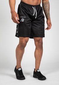 Zwarte sportieve shorts met een mesh structuur, elastische tailleband, witte trekkoorden en gedrukte graphics aan de zijkant, gedragen met zwarte sneakers.
