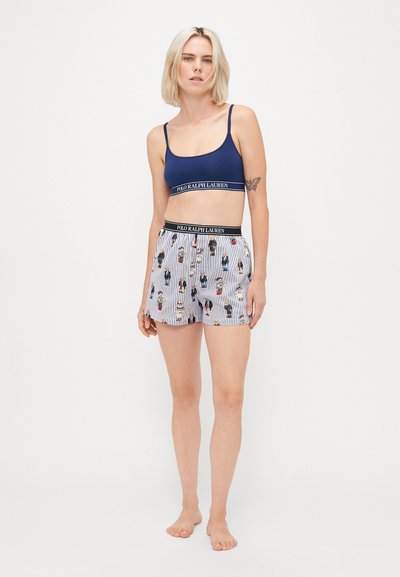 Žena s krátkymi blond vlasmi, oblečená v tmavomodrej bralette a bielych pruhovaných šortkách so vzorom medveďov a pásikmi s logom Polo Ralph Lauren.