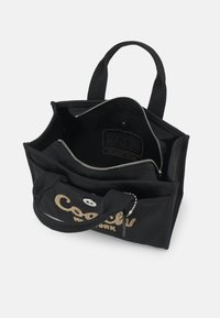 Black canvas tote bag with top zip closure, two interior compartments, and decorative gold lettering. Features sturdy handles and metal accents.