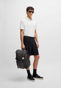 White polo shirt, tailored black shorts, and black sneakers with socks. Carrying a gray backpack featuring multiple compartments and zippers.