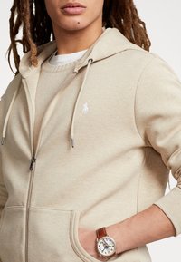 Beige zip-up hoodie with a front pocket, white logo on the chest, and drawstrings. Worn over a light sweater with a textured finish.
