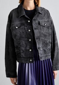 Svart denimjacka med en kortad design, frontknappar, två bröstfickor och synlig sömnad. Bärs över en veckad lila kjol.
