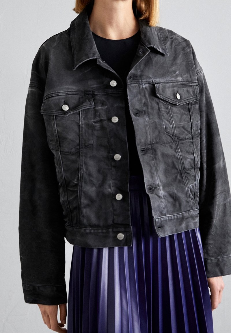Svart denimjacka med en kortad design, frontknappar, två bröstfickor och synlig sömnad. Bärs över en veckad lila kjol.