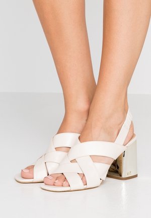 Beige åbne sandaler med krydsede stropper, en polstret hæl og en metallisk blokhæl. Glat læderfinish med en flad sål.