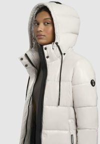 Puffjakke i off-white med en stor hætte, lynlås lukning og sorte detaljer. Tekstureret, quiltet design med mærkebaserede hardware detaljer.
