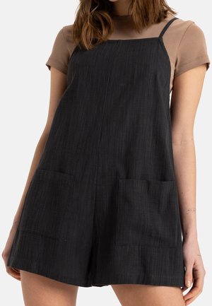 Woman wearing black sleeveless romper with front pockets over a short-sleeve beige top, standing against a plain background.