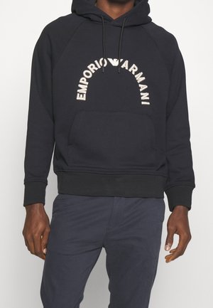 Mężczyzna w czarnej bluzie z kapturem Emporio Armani z przednią kieszenią i logo w formie łuku na piersi.