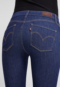 Jeans de mezclilla azul oscuro con un diseño ajustado, que presentan una cintura alta, bolsillos traseros curvados y costuras doradas en contraste.