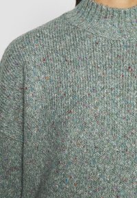 Suéter tejido en verde claro con un acabado texturizado. Presenta motas coloridas en varios tonos, cuello alto y un ajuste holgado.