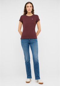 Femme debout les mains derrière le dos, portant un T-shirt à manches courtes bordeaux, un jean bleu et des baskets blanches avec des rayures noires.
