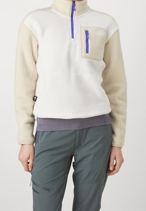 Person wearing a beige and white fleece pullover with a blue zipper pocket and dark green pants.