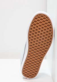 Semelle d'une basket blanche avec une semelle extérieure en caoutchouc marron à motif gaufré, sur fond d'angle blanc.