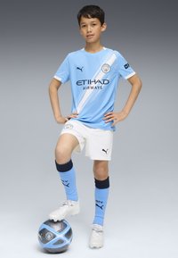 Equipación de fútbol de color azul claro del Manchester City con detalles en blanco, que presenta un logo y texto del patrocinador. El jugador está de pie con un pie sobre un balón de fútbol azul y negro.