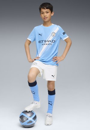 Vaaleansininen Manchester City -jalkapallopuku vaaleanvalkoisilla yksityiskohdilla, jossa on logo ja sponsoriteksti. Pelaaja seisoo jalka sinimustassa jalkapallossa.