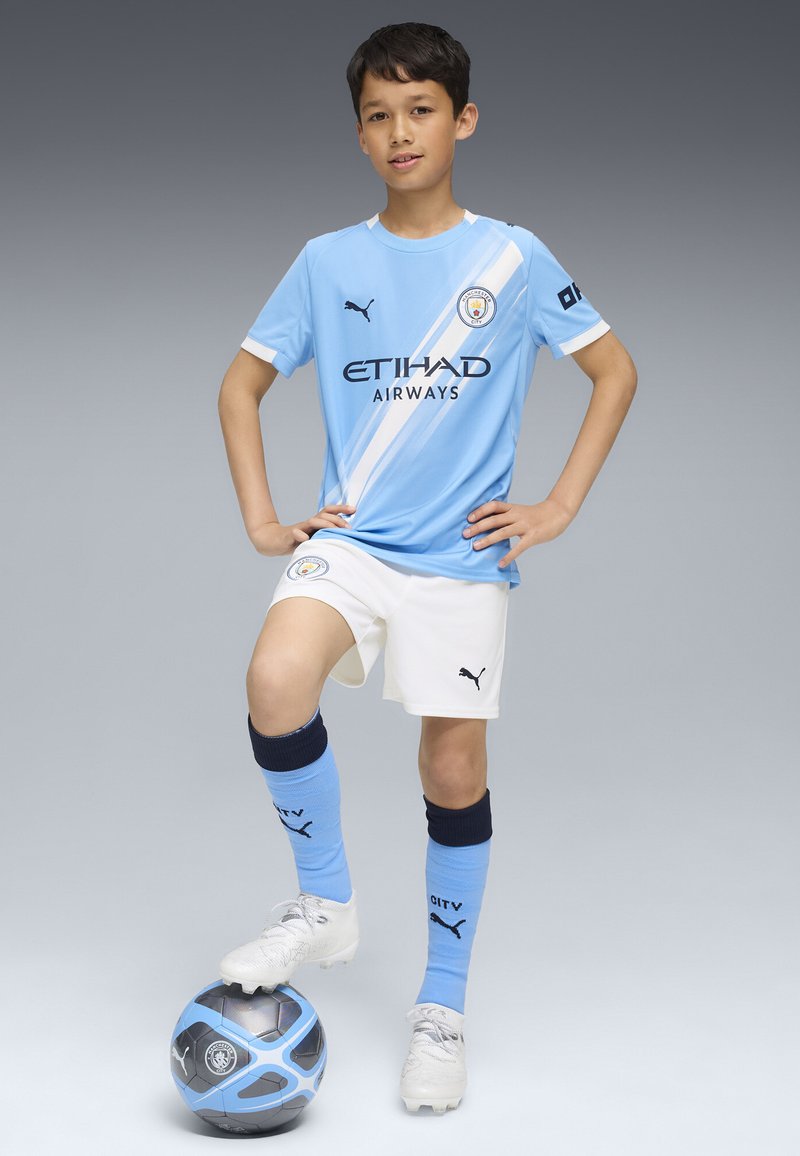 Equipación de fútbol de color azul claro del Manchester City con detalles en blanco, que presenta un logo y texto del patrocinador. El jugador está de pie con un pie sobre un balón de fútbol azul y negro.