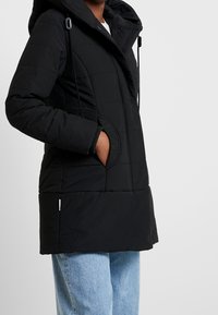 Svart vadderad pufferjacka med huva, som har justerbara snören, texturerade sidofickor och ribbade detaljer vid ärmsluten.