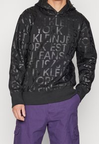 Black hoodie featuring a textured design with reflective black lettering, ribbed cuffs, and a drawstring hood. Paired with purple pants.