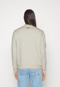 Beige sweatshirt med rund halsringning och långa ärmar. Har en liten svart "EA7"-logotyp på baksidan. Slät textur, avslappnad passform.