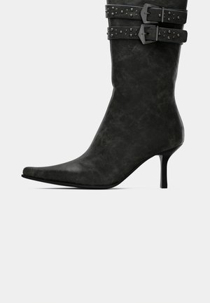 Zwarte leren laars tot halverwege de kuit met een puntige neus, slanke hoge hak en twee gesp met studs rond de bovenkant.