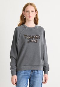 Felpa grigia con logo "TOMMY JEANS". Presenta un collo rotondo, maniche raglan e polsini a coste. Indossata con jeans in denim blu chiaro.