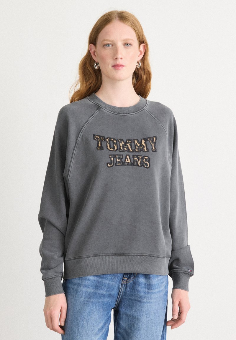 Felpa grigia con logo "TOMMY JEANS". Presenta un collo rotondo, maniche raglan e polsini a coste. Indossata con jeans in denim blu chiaro.