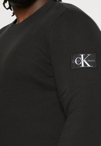 Chemise noire à manches longues en tissu texturé. Présente un patch logo Calvin Klein Jeans sur la manche droite. Design simple et ajusté.