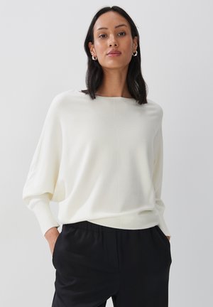 Off-white, long-sleeve knit sweater with a boat neckline, relaxed fit, and ribbed cuffs, worn with black elastic waist trousers.
