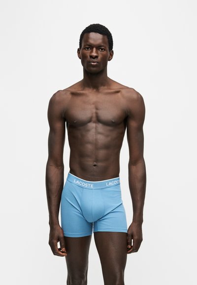 Homme torse nu debout, portant un boxer bleu clair Lacoste, face à l'avant sur fond blanc uni.