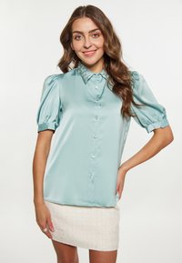 Blusa di raso azzurro chiaro con maniche corte a palloncino, colletto di perline e chiusura frontale con bottoni, abbinata a una gonna bianca strutturata.