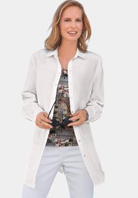 White button-up shirt with long sleeves, layered over a multicolored patterned top. Model holds sunglasses, against a neutral background.