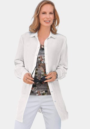 White button-up shirt with long sleeves, layered over a multicolored patterned top. Model holds sunglasses, against a neutral background.
