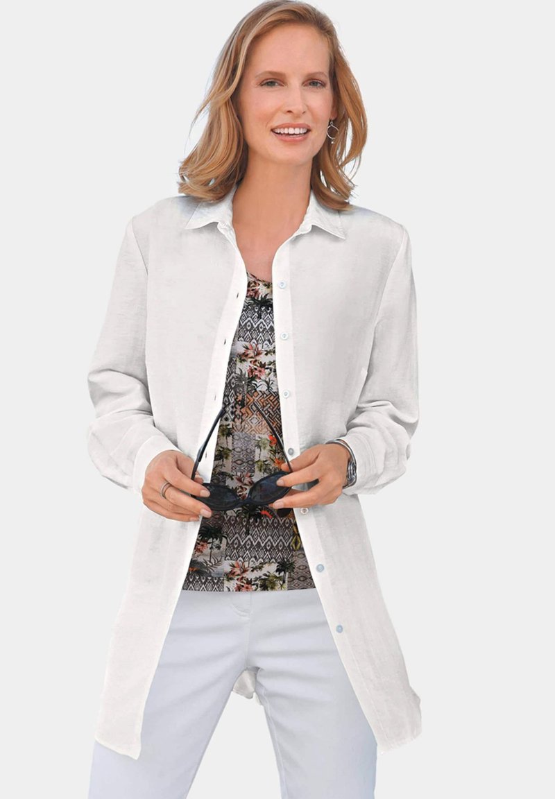 White button-up shirt with long sleeves, layered over a multicolored patterned top. Model holds sunglasses, against a neutral background.