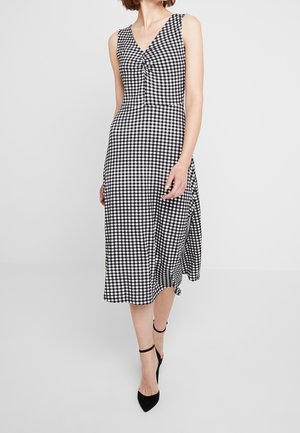 Kvinde iklædt sort-hvid ternet ærmeløs midi-kjole med V-hals og sorte ankelrem hæle, stående på grå baggrund.