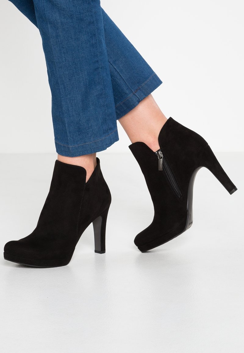 Bottines en daim noir à talons hauts avec fermeture éclair latérale, portées avec un jean denim bleu raccourci sur un sol clair.